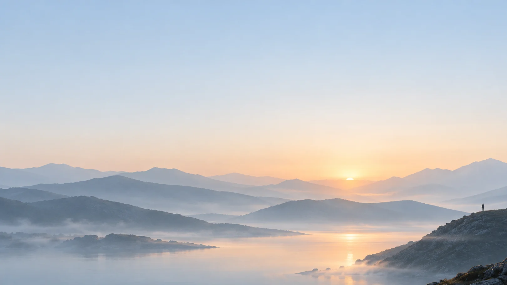 Amanecer sobre colinas en capas con niebla y un lago quieto; una figura diminuta mira desde una roca.
